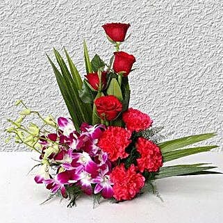 Cane basket arrangement of 4 purple orchids, 4 dark pink carnations, 4 red roses and arica palm leaves